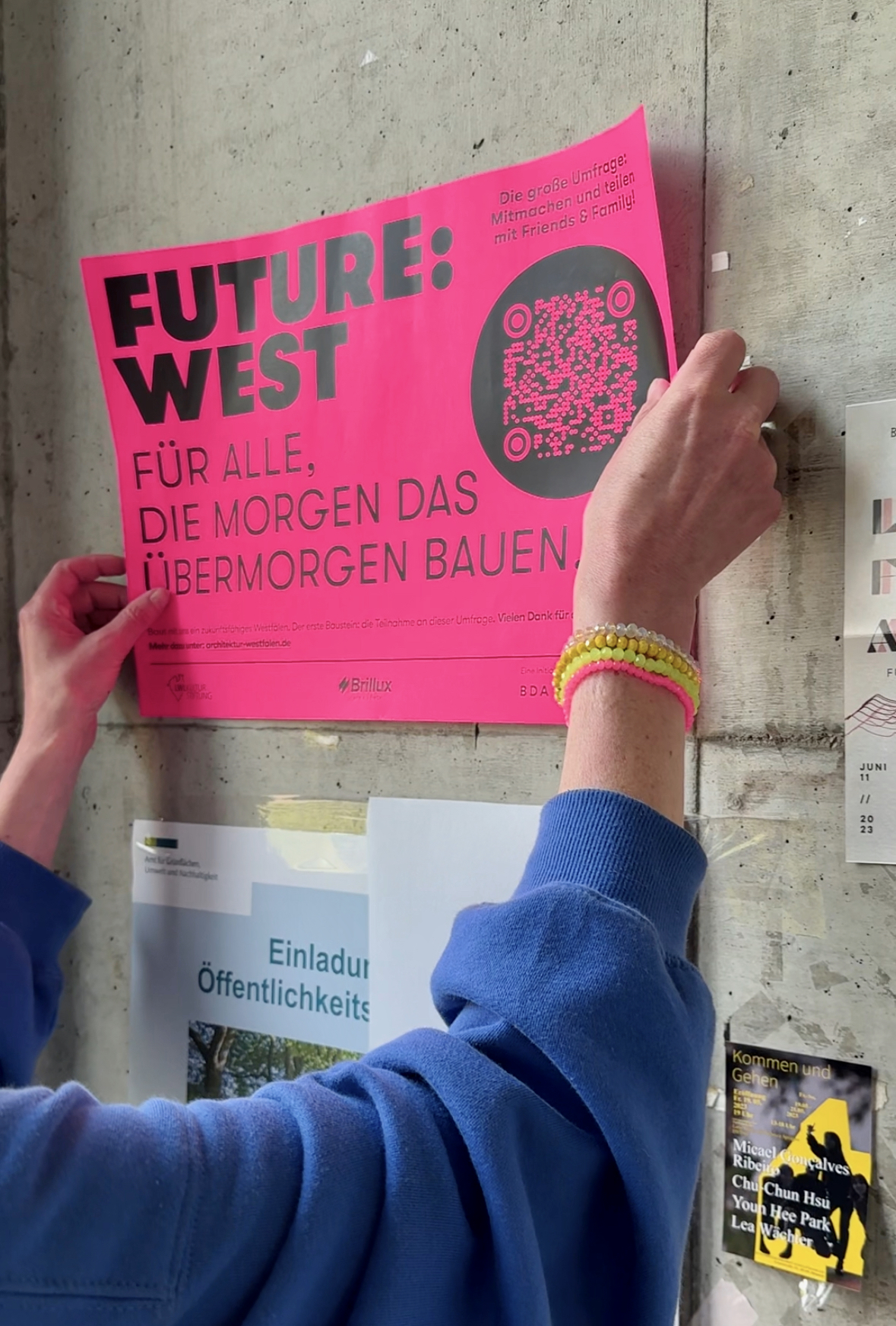 Eine Frau (man sieht nur die Arme) hängt ein Plakat auf, das für die Teilnahme an der Umfrage FUTURE:WEST wirbt.